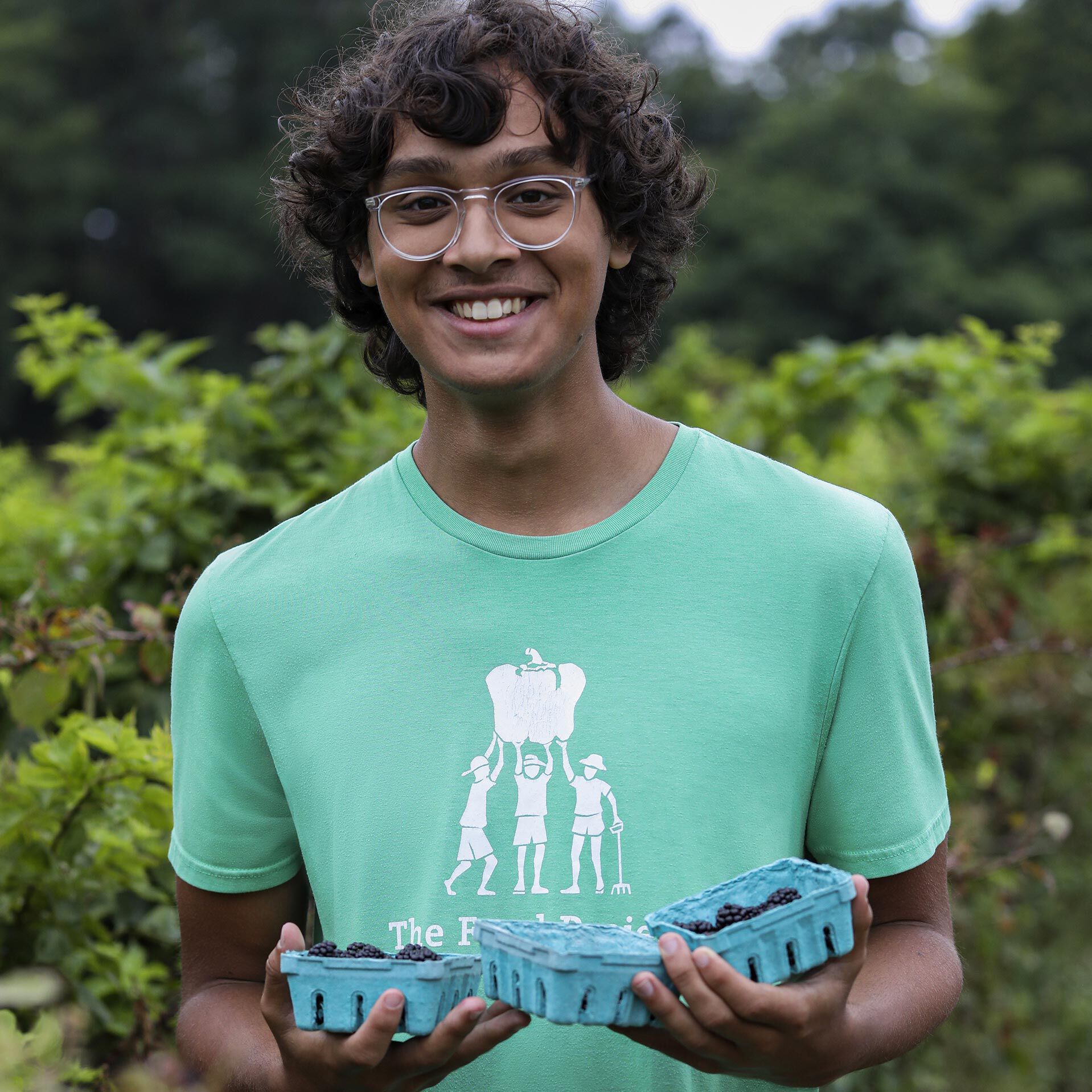 A medium-skinned, male-presenting teen holding several containers of blackberries he’s harvested himself as a farm volunteer.
