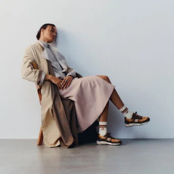 A woman sitting on a wooden chair with her legs crossed wearing a pair of New Balance Made in UK sneakers in a minimalist room with a white wall and concrete floor