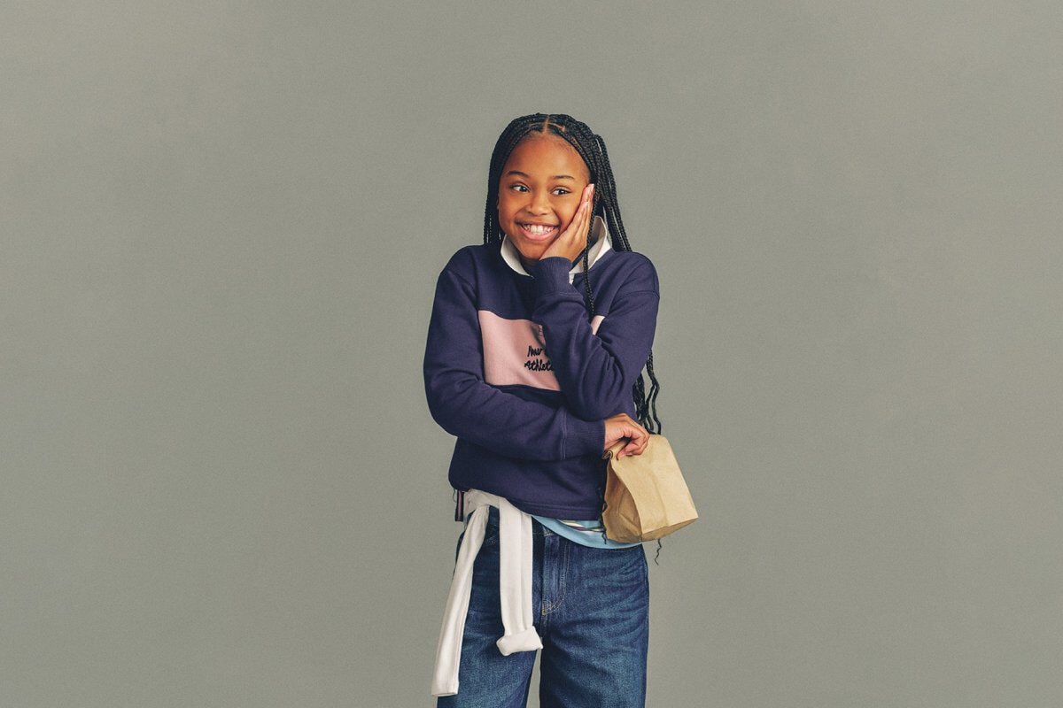 The child is wearing a light gray T-shirt layered under a soft pink zip-up hoodie with a white New Balance logo on the chest.
