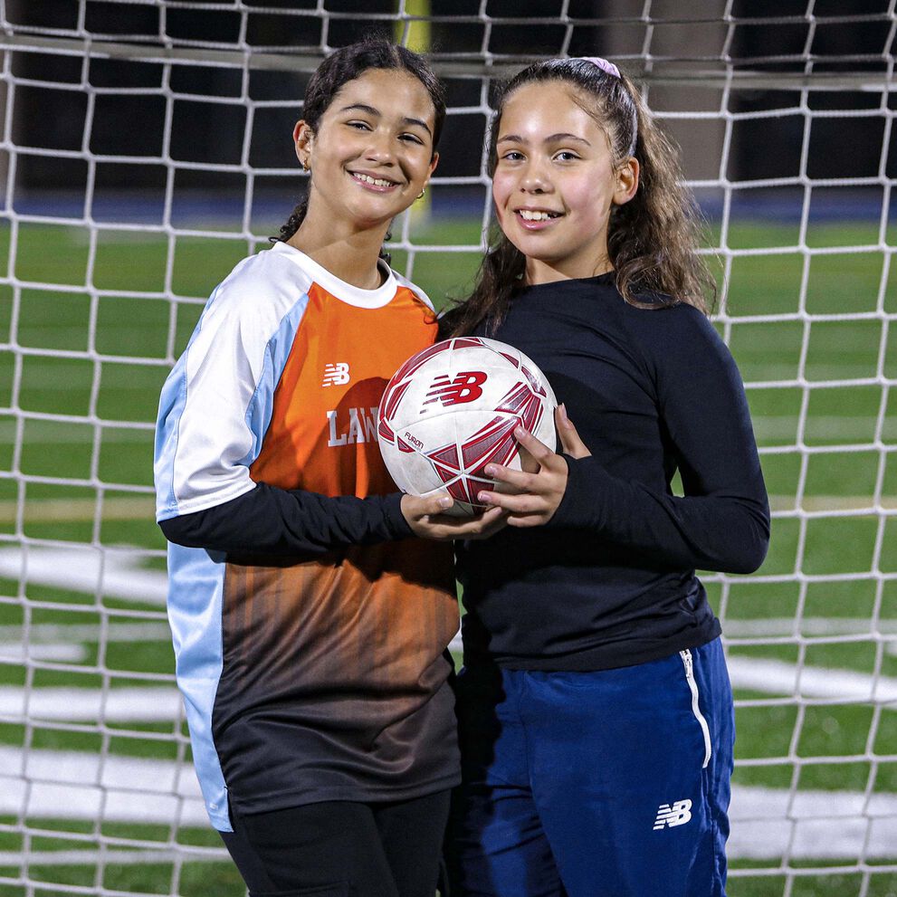 Two smiling teen girls posing with a soccer ball in front of a goal.